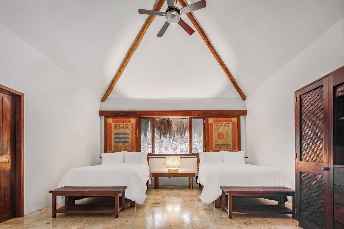 A room with two beds, a ceiling fan, a window with a view, and a wooden door.