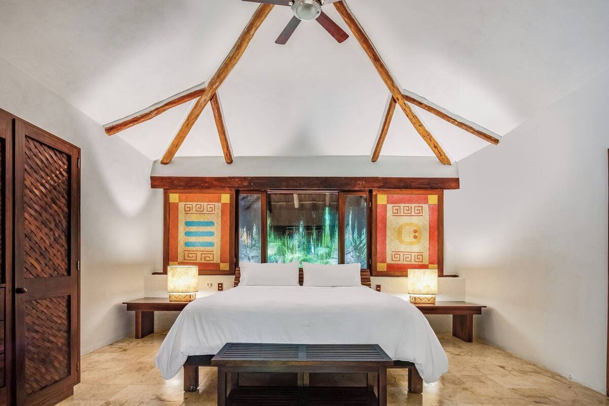 A bedroom with a bed, two bedside tables, a ceiling fan, and a window with a view of greenery.