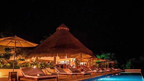 A nighttime pool area with lounge chairs and umbrellas, a thatched-roof structure, and a bar area visible in the background. - The Explorean Selva Maya Kohunlich (Kohunlich)