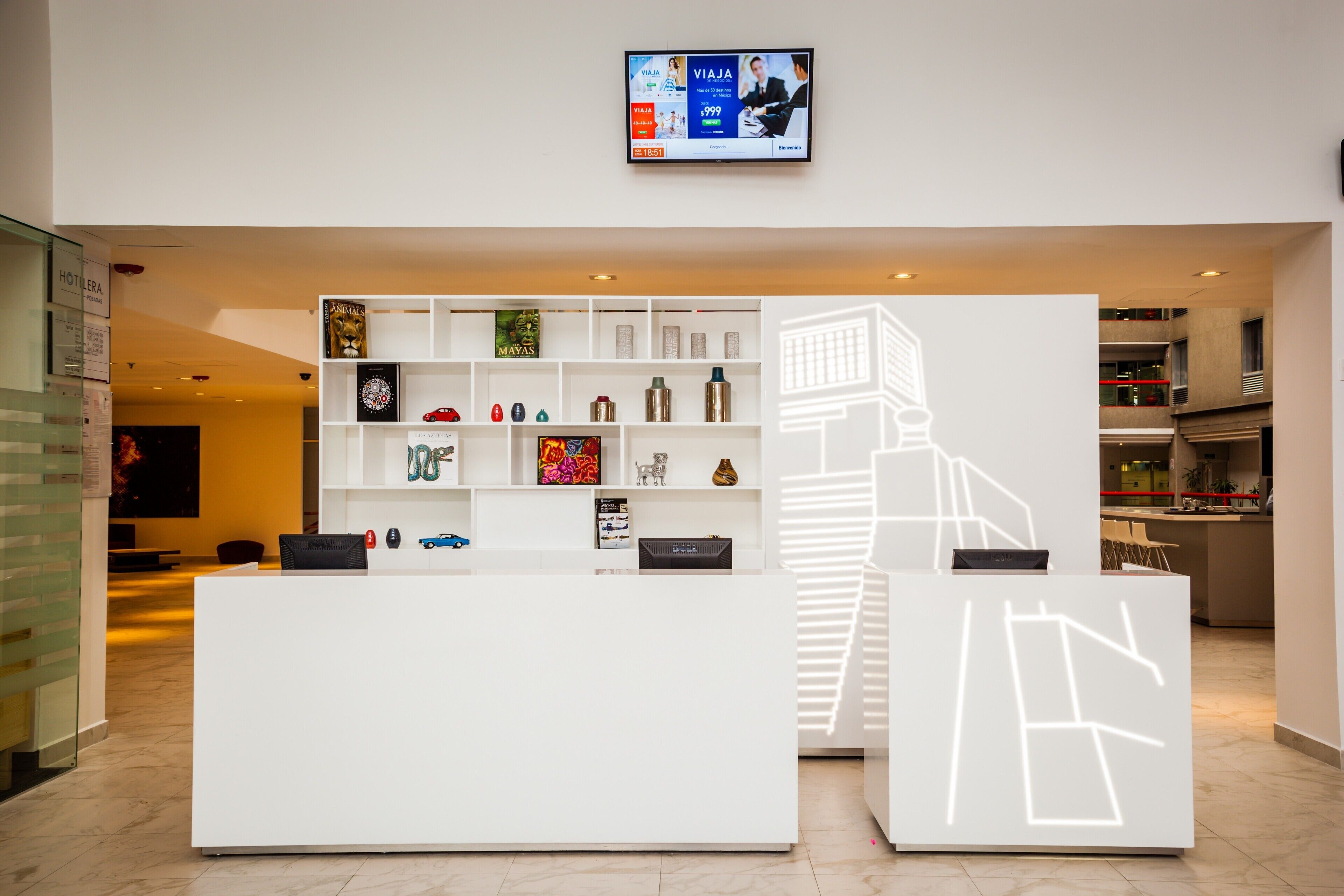Una moderna hall d'albergo con una reception, uno schermo digitale che mostra una pubblicità turistica e una libreria con diversi oggetti decorativi.