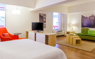 Una habitación de hotel con cama, televisor, escritorio, sofá verde y sillón rojo.
