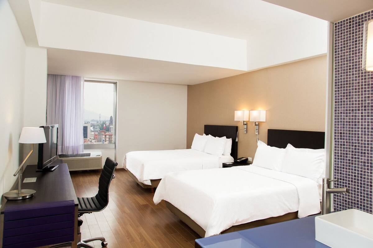 A hotel room with two beds, a desk with a lamp, a chair, a window with a view, and a bathroom with a tiled wall.