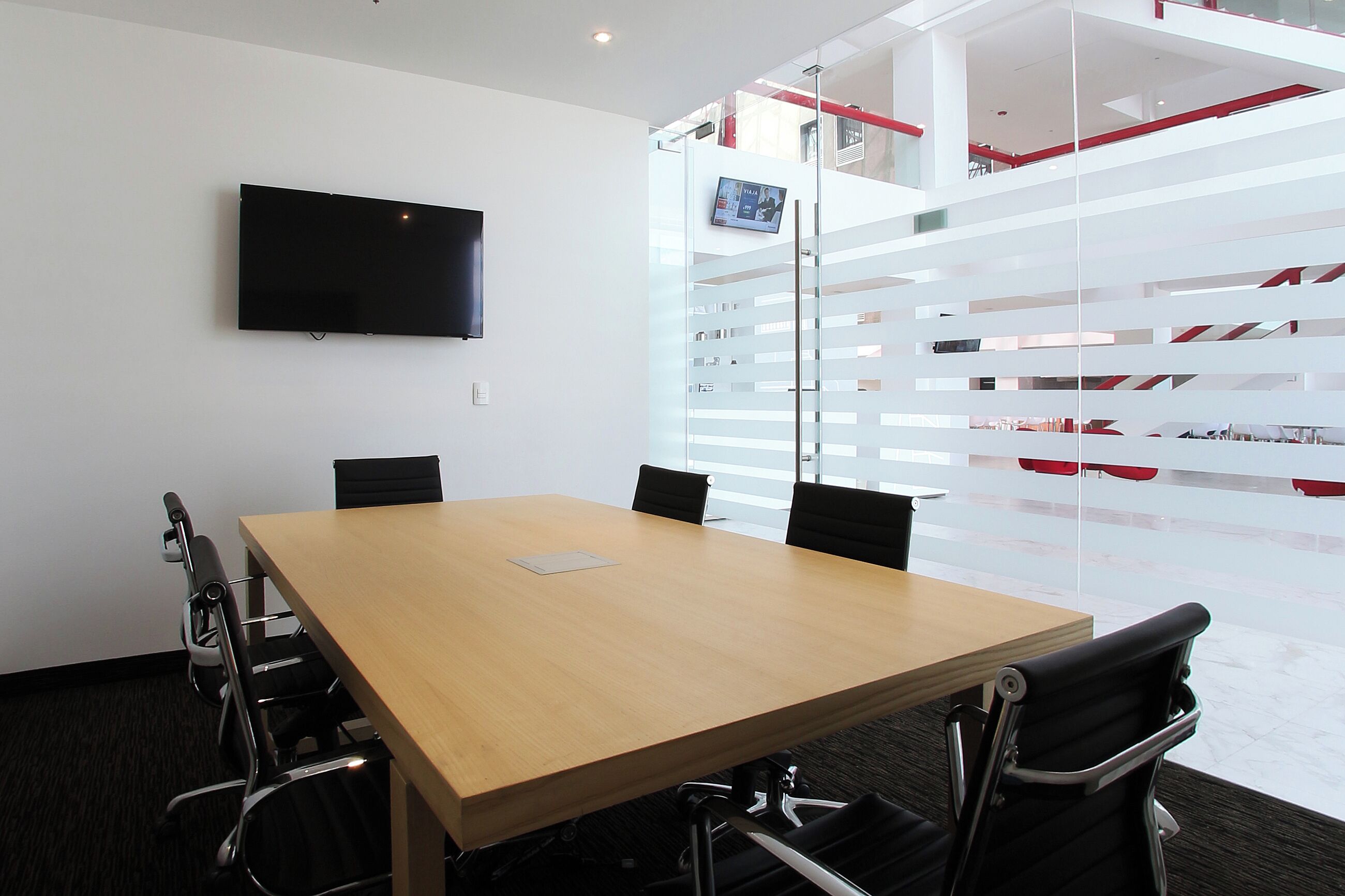 Un salón de conferencias con una gran mesa, cuatro sillas, un televisor de pantalla plana y un panel de vidrio con detalles rojos.