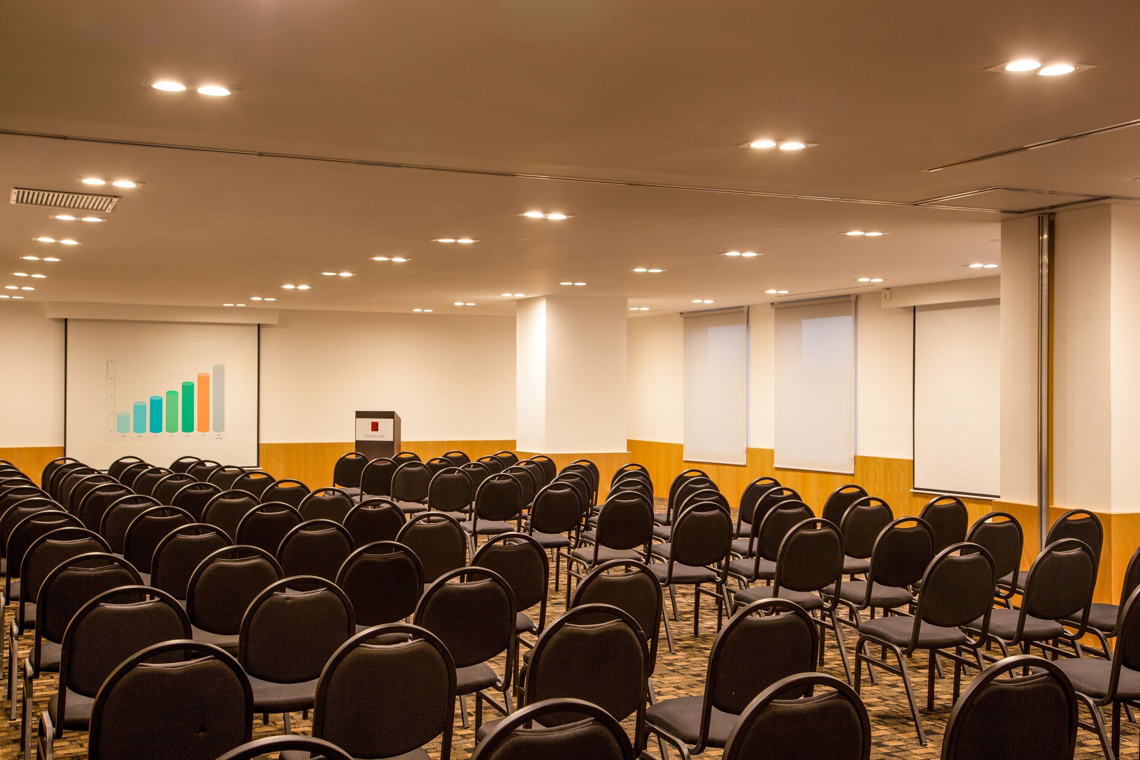 A conference room with rows of chairs, a presentation screen, and a carpeted floor.