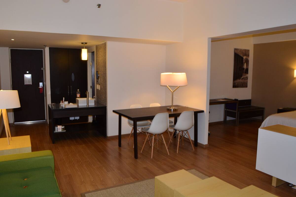 A hotel room with a wooden floor, a dining table with chairs, a bed, a desk, a lamp, and a green sofa.