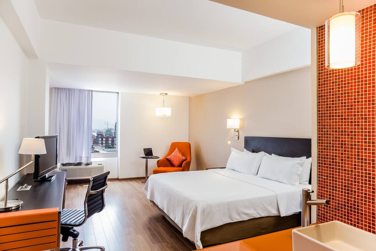 A hotel room with a bed, desk, chair, and a view of the city.