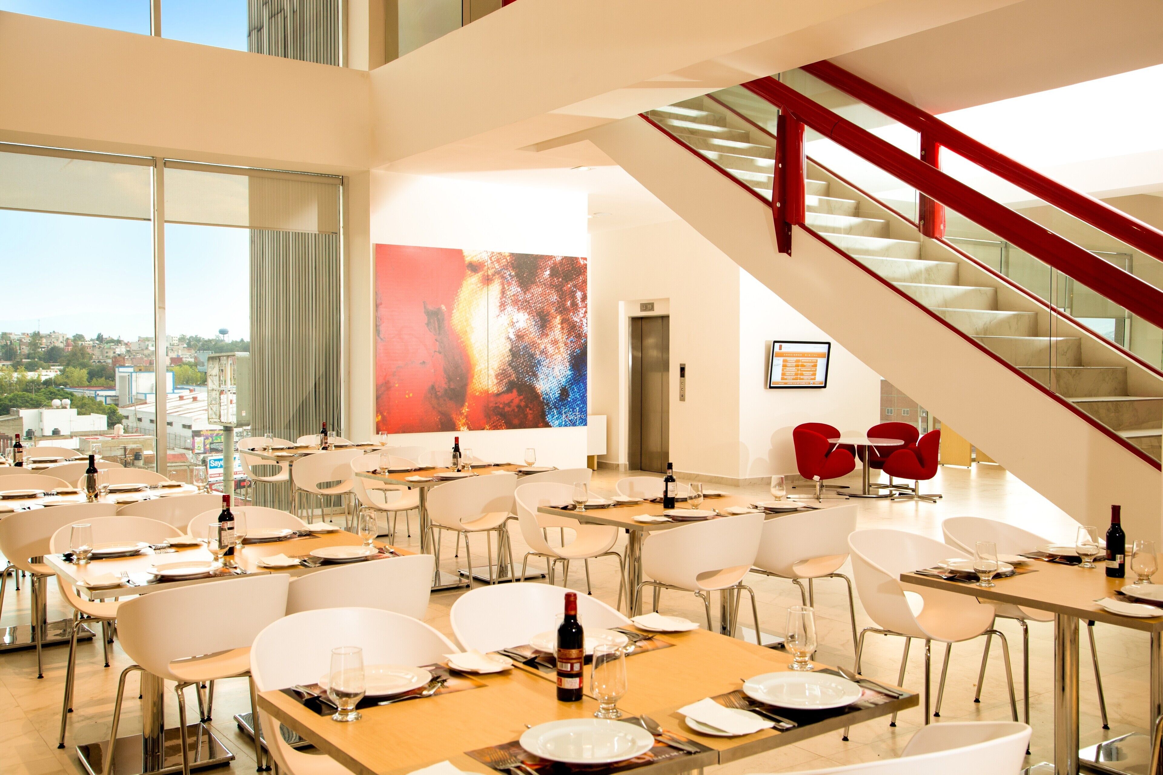 Un comedor moderno con varias mesas y sillas, una escalera y un amplio ventanal con vistas a la ciudad.