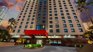 A high-rise hotel with a modern design, featuring a central entrance with a red canopy, multiple floors, and a surrounding landscaped area with palm trees. - CHN Hotel Monterrey Centro, Trademark by Wyndham (Monterrey)