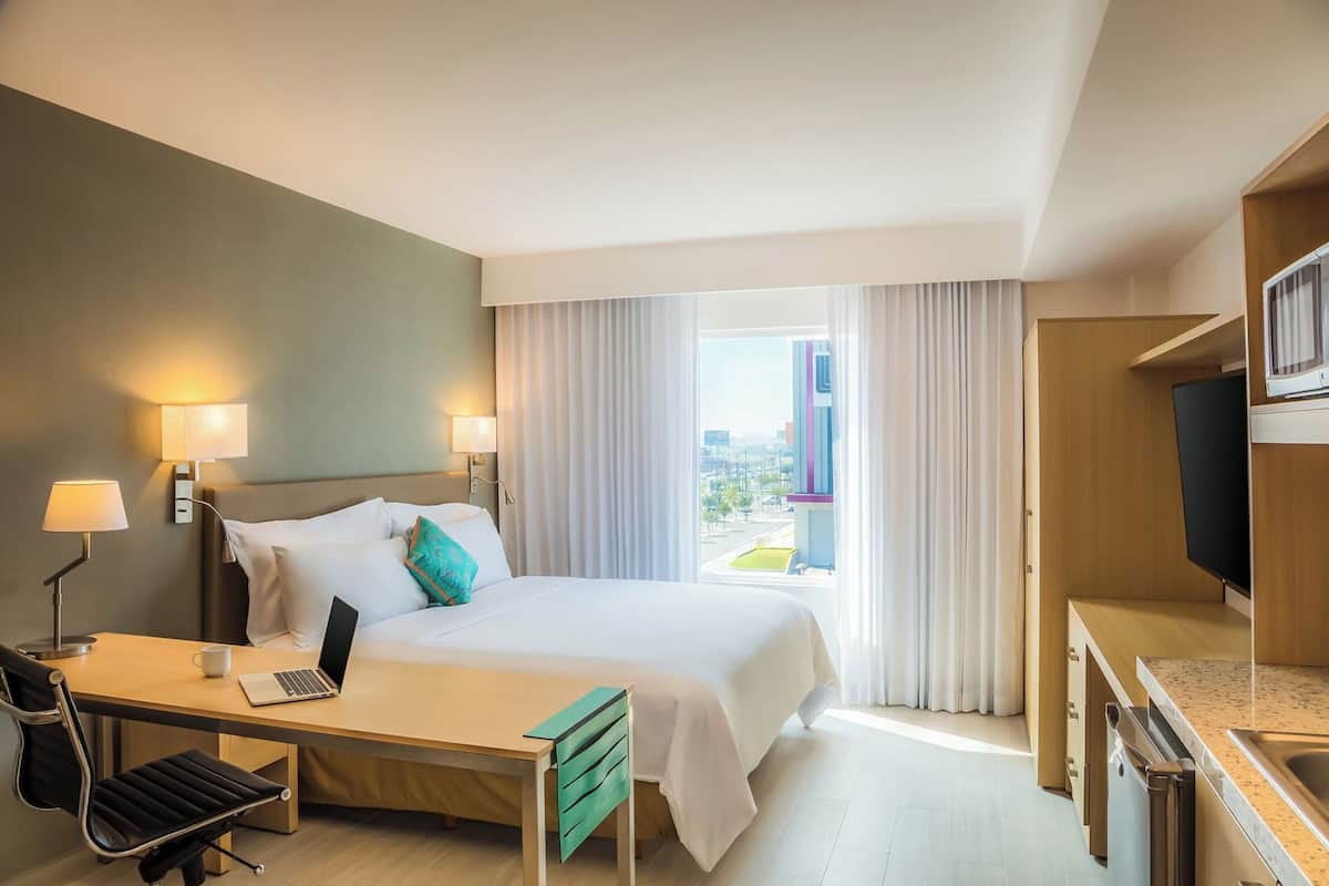 A hotel room with a bed, desk, chair, TV, and a view of the city.