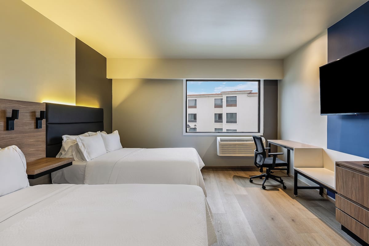 A hotel room with two beds, a desk, a chair, a TV, and a window with a view of buildings.