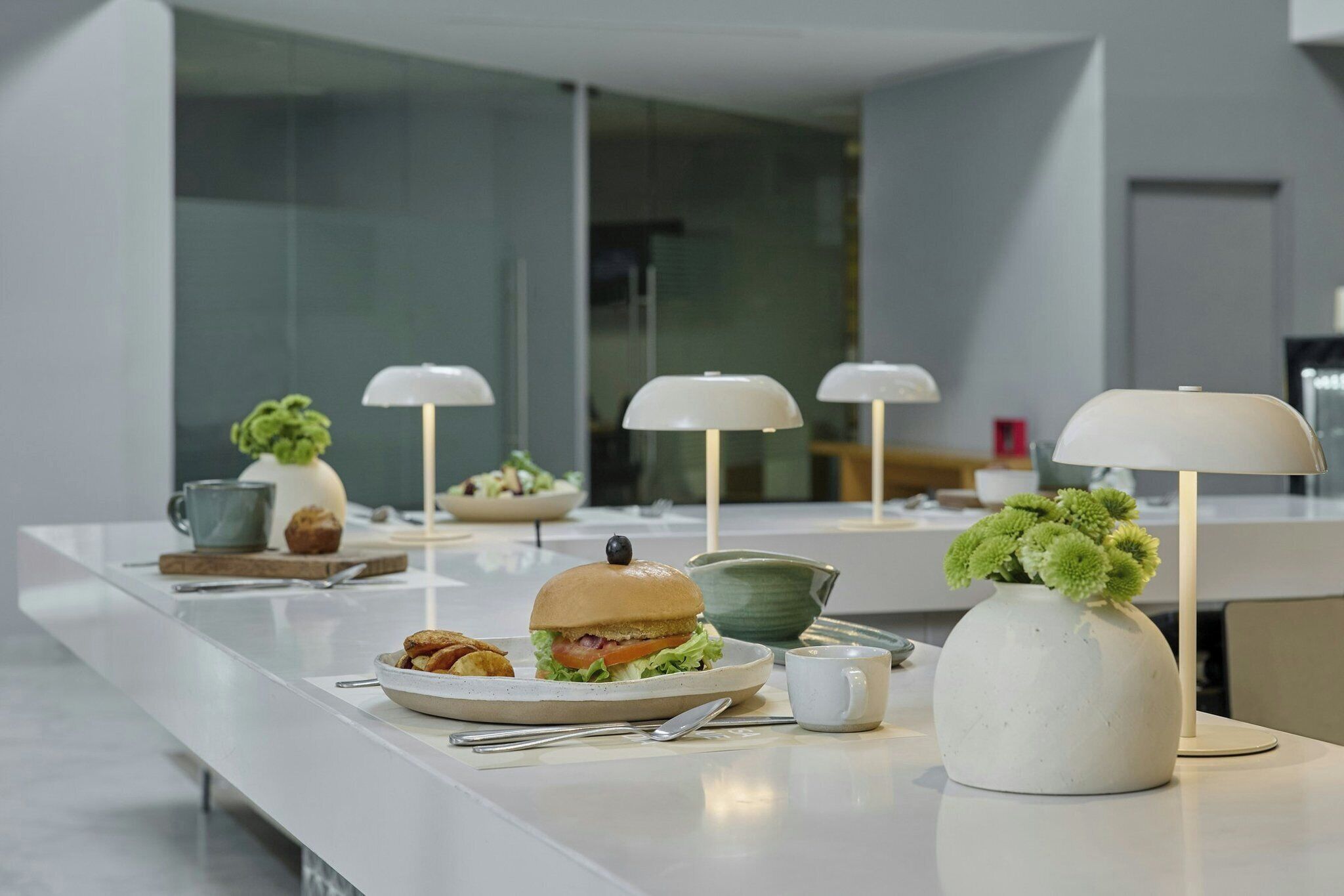Una cocina moderna con una encimera larga, un sándwich, un tazón de ensalada, una taza de café y un florero con flores verdes.