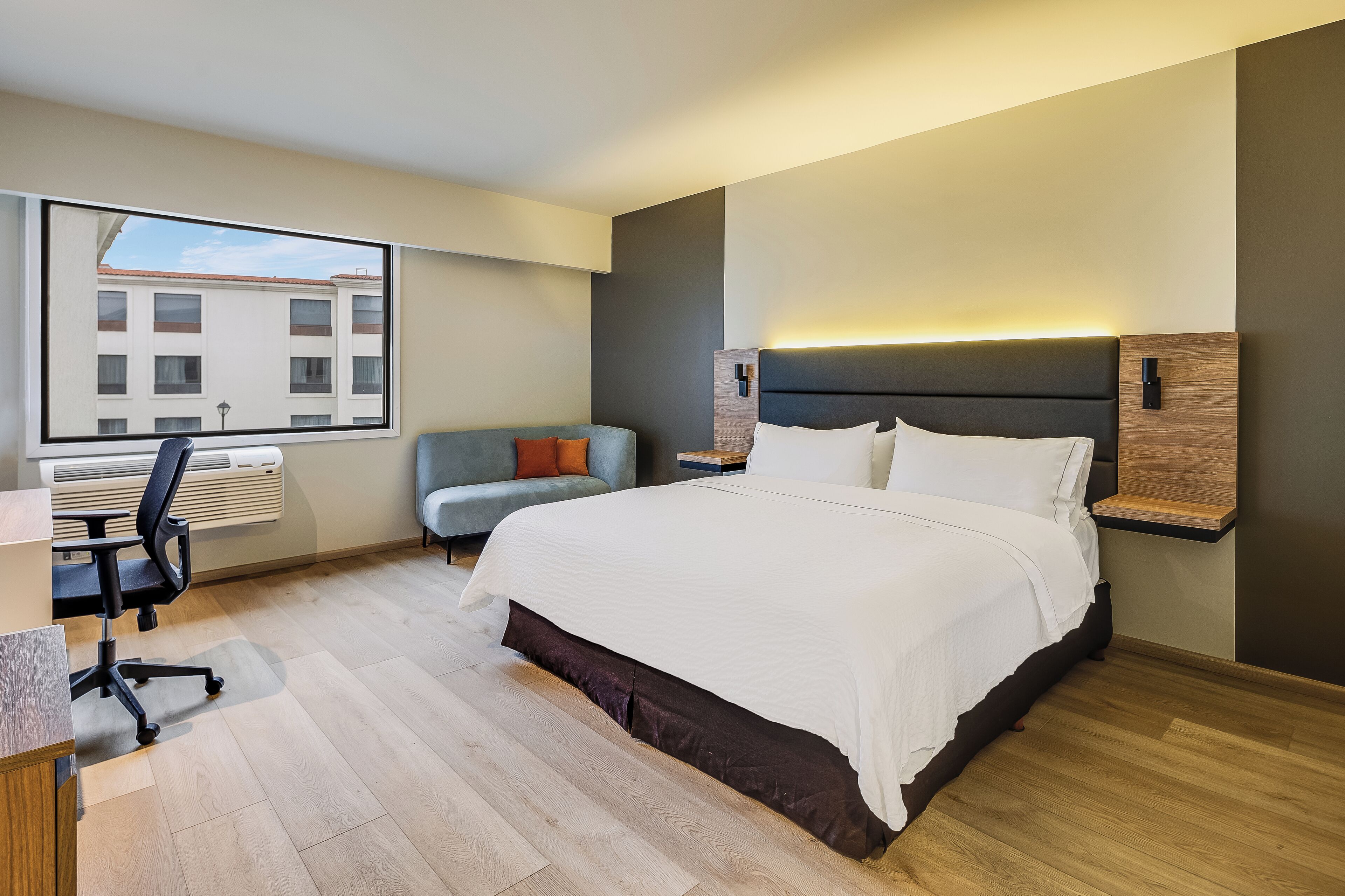 A modern hotel room with a large bed, a desk, a chair, and a window with a view of buildings.
