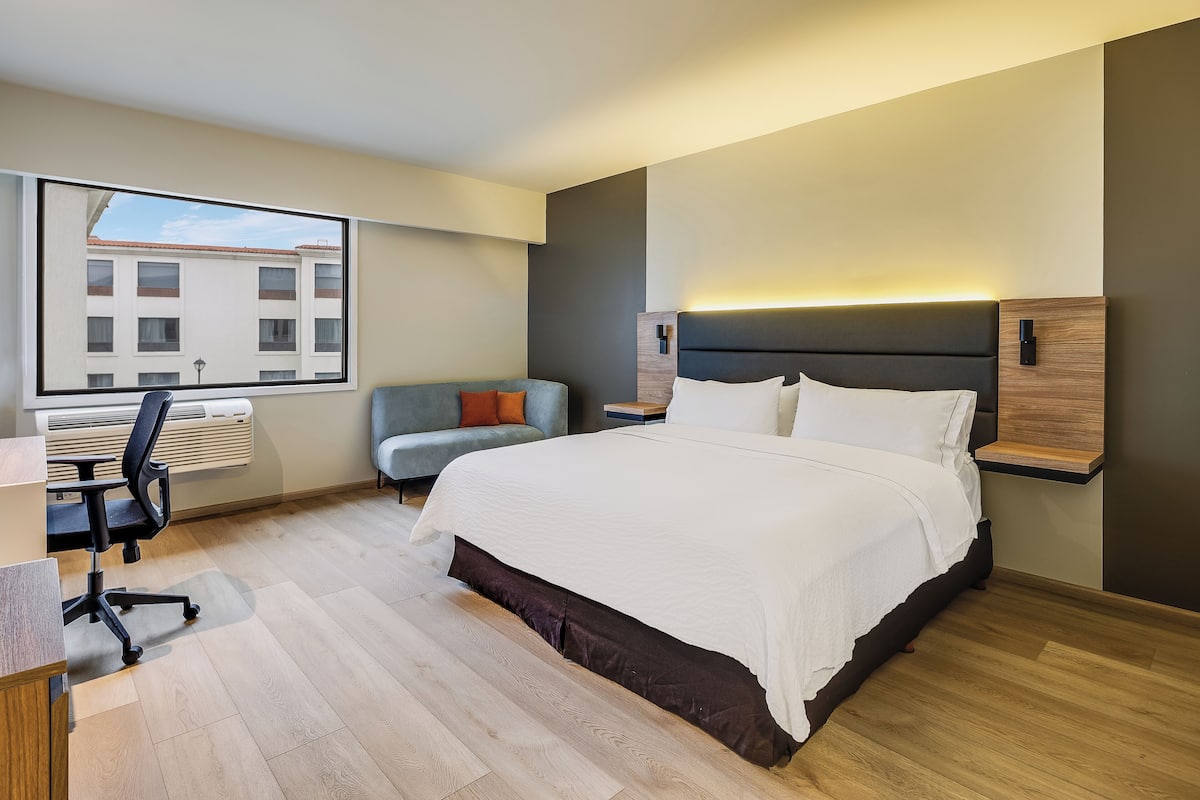A modern hotel room with a large bed, a desk, a chair, and a window with a view of buildings.