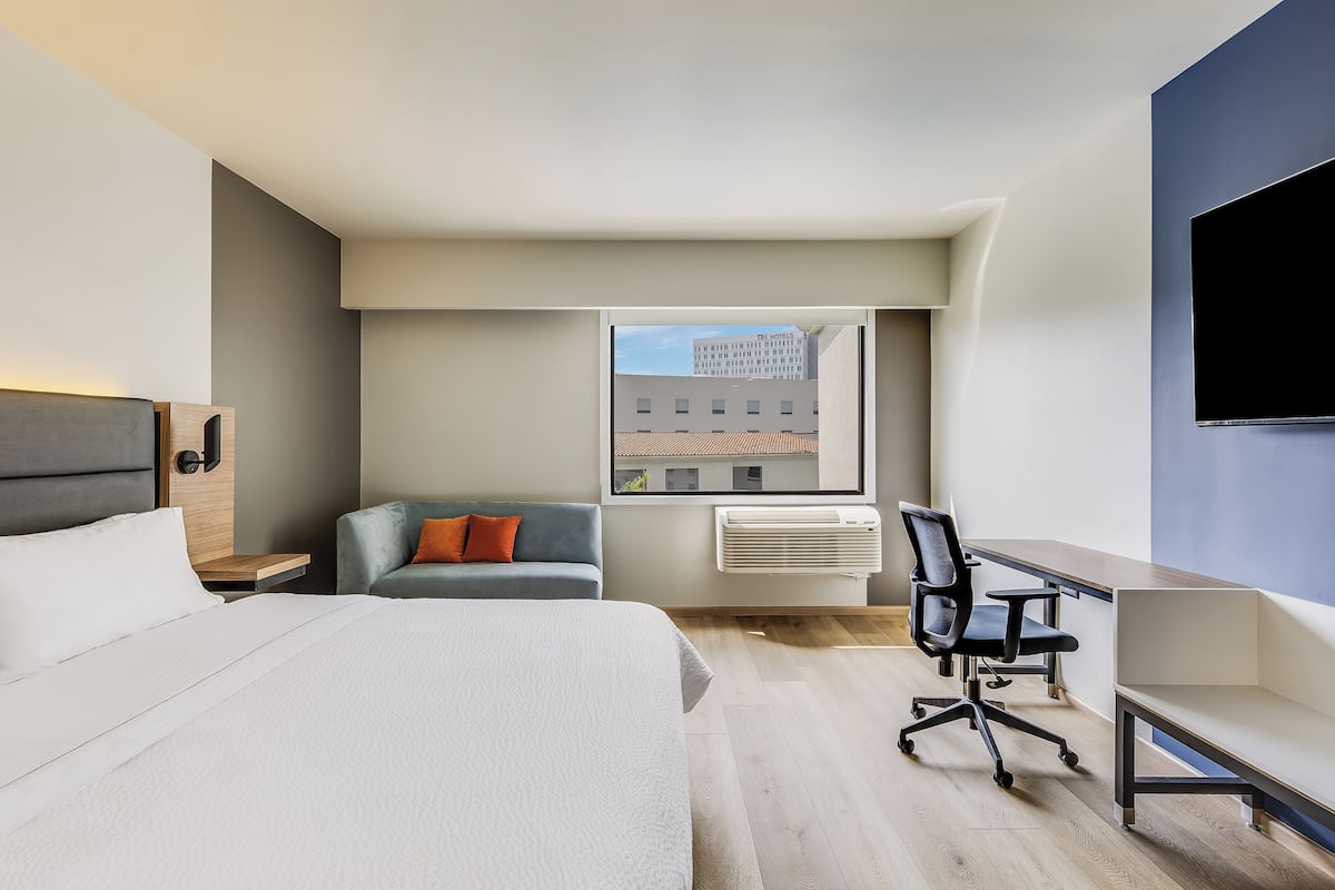 A modern hotel room with a bed, a desk, a chair, a sofa, and a large window with a city view.