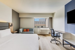 A modern hotel room with a bed, a desk, a chair, a sofa, and a large window with a city view.