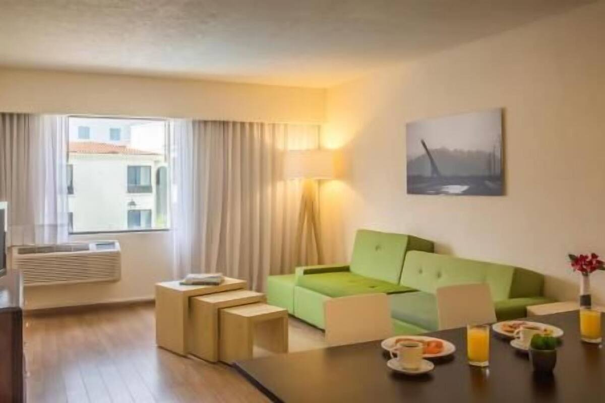 A hotel room with a green sofa, a dining table with breakfast, a TV, and a window with curtains.