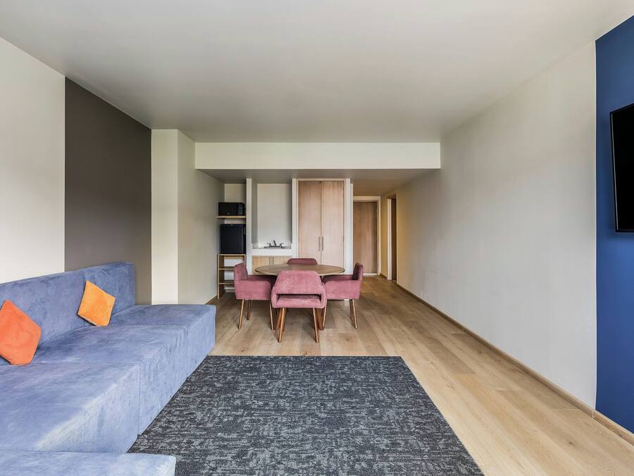 A modern living room with a blue sofa, a dining table with pink chairs, and a kitchen area in the background.