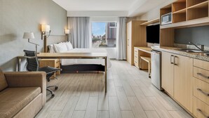 A hotel room with a bed, desk, chair, kitchenette, and a view of the city.