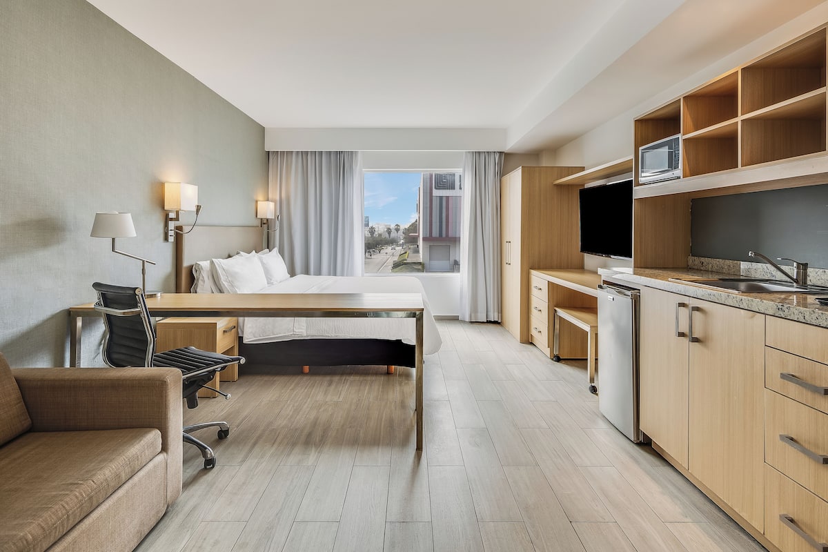 A hotel room with a bed, desk, chair, kitchenette, and a view of the city.