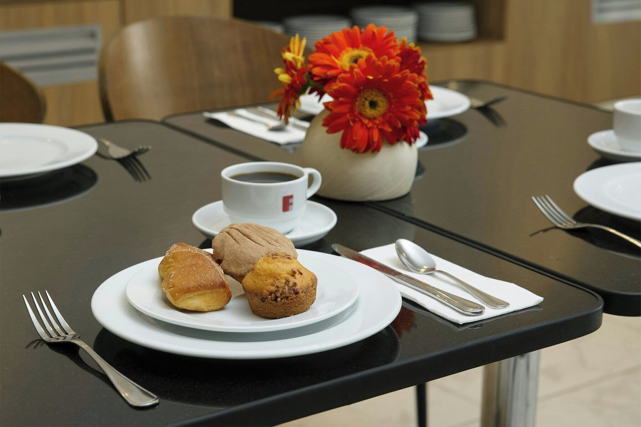Una mesa dispuesta con una taza de café, un plato de muffins y un florero con flores.