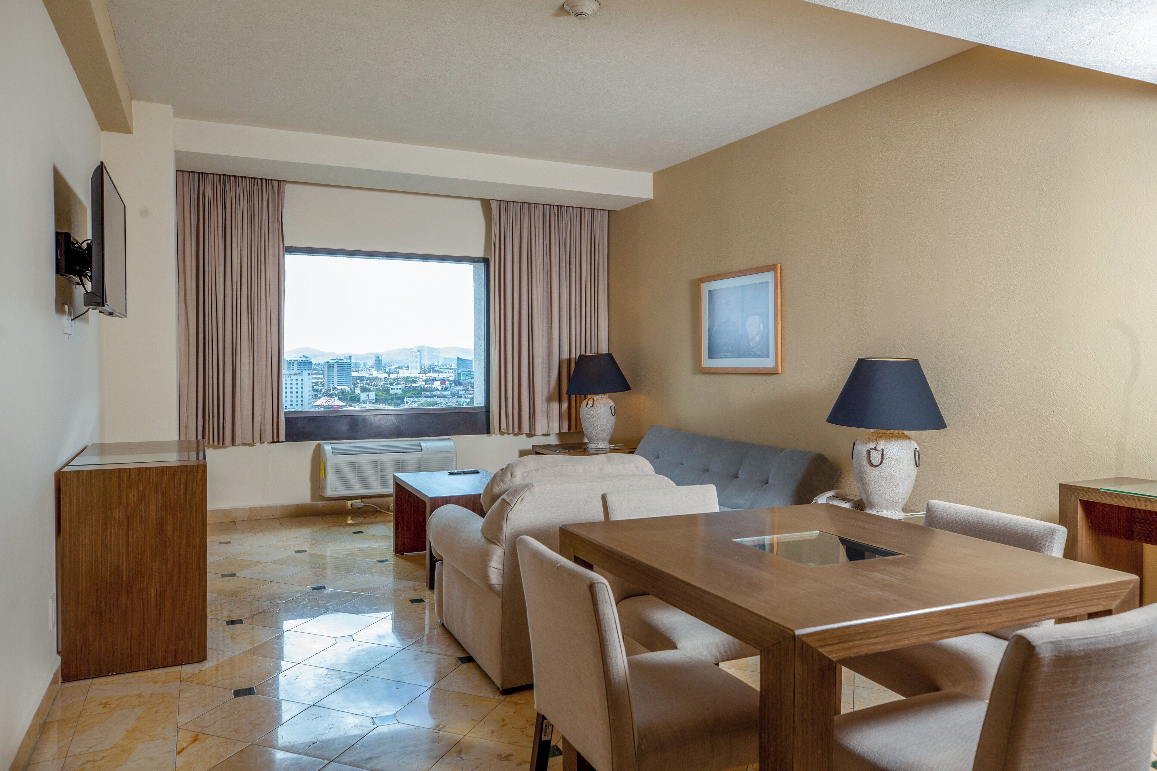Quarto de hotel com área de jantar, sofá, duas luminárias, televisão e uma grande janela com cortinas.