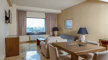 Quarto de hotel com área de jantar, sofá, duas luminárias, televisão e uma grande janela com cortinas.