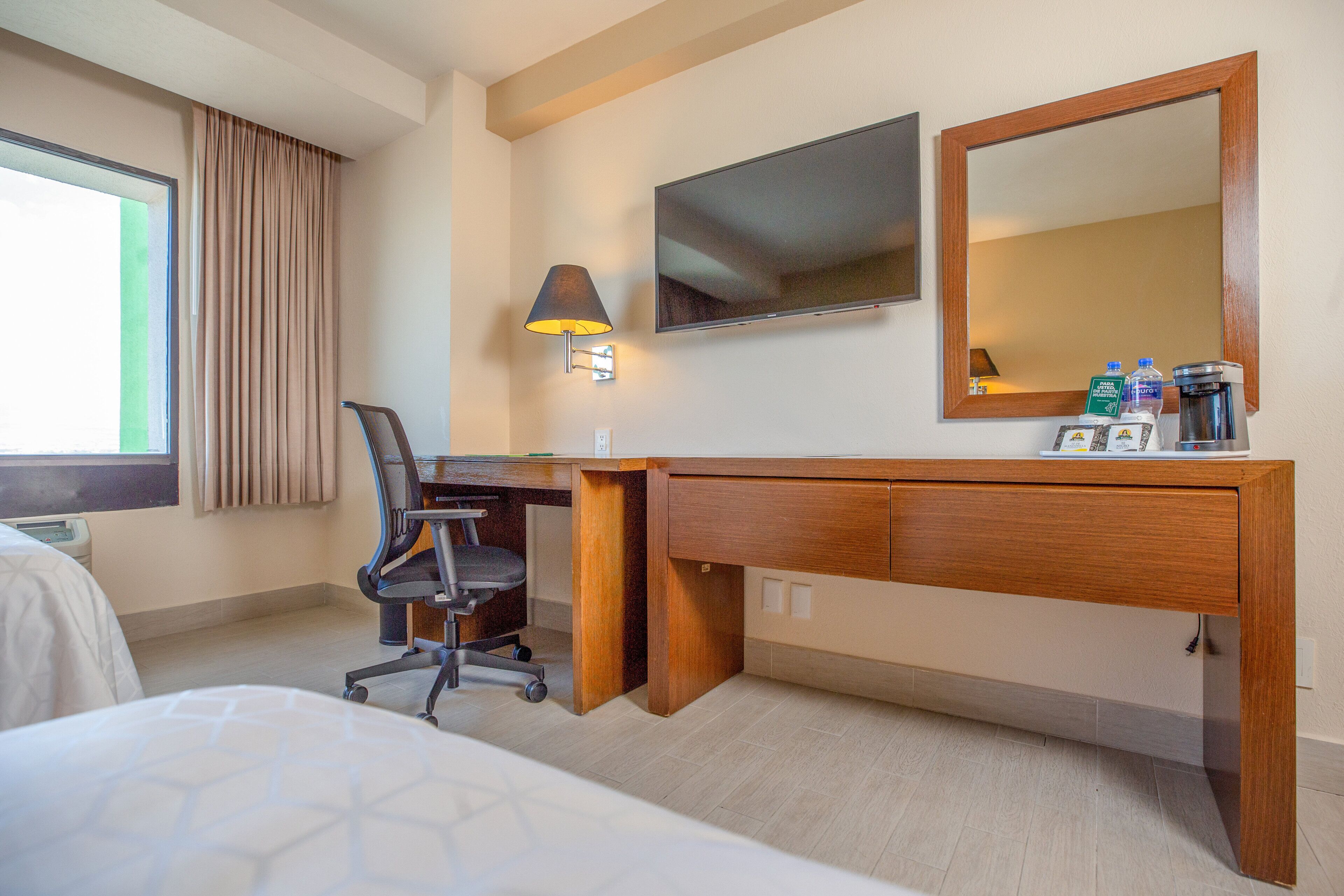 Habitación de hotel con cama, escritorio, silla, televisor, lámpara, espejo y ventana con cortinas.