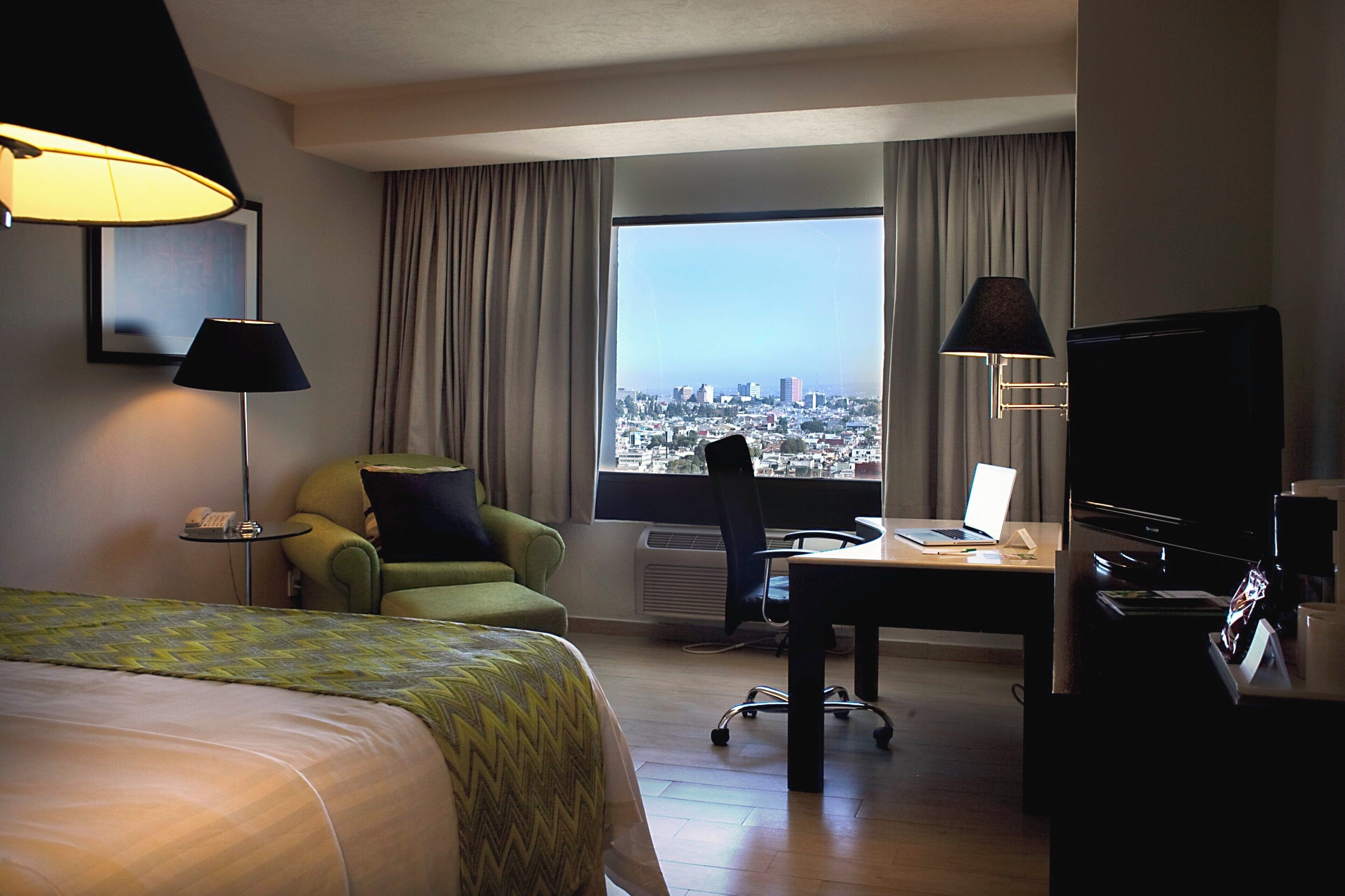 Habitación de hotel con cama, escritorio con una computadora portátil, una silla, televisión y vista a la ciudad.