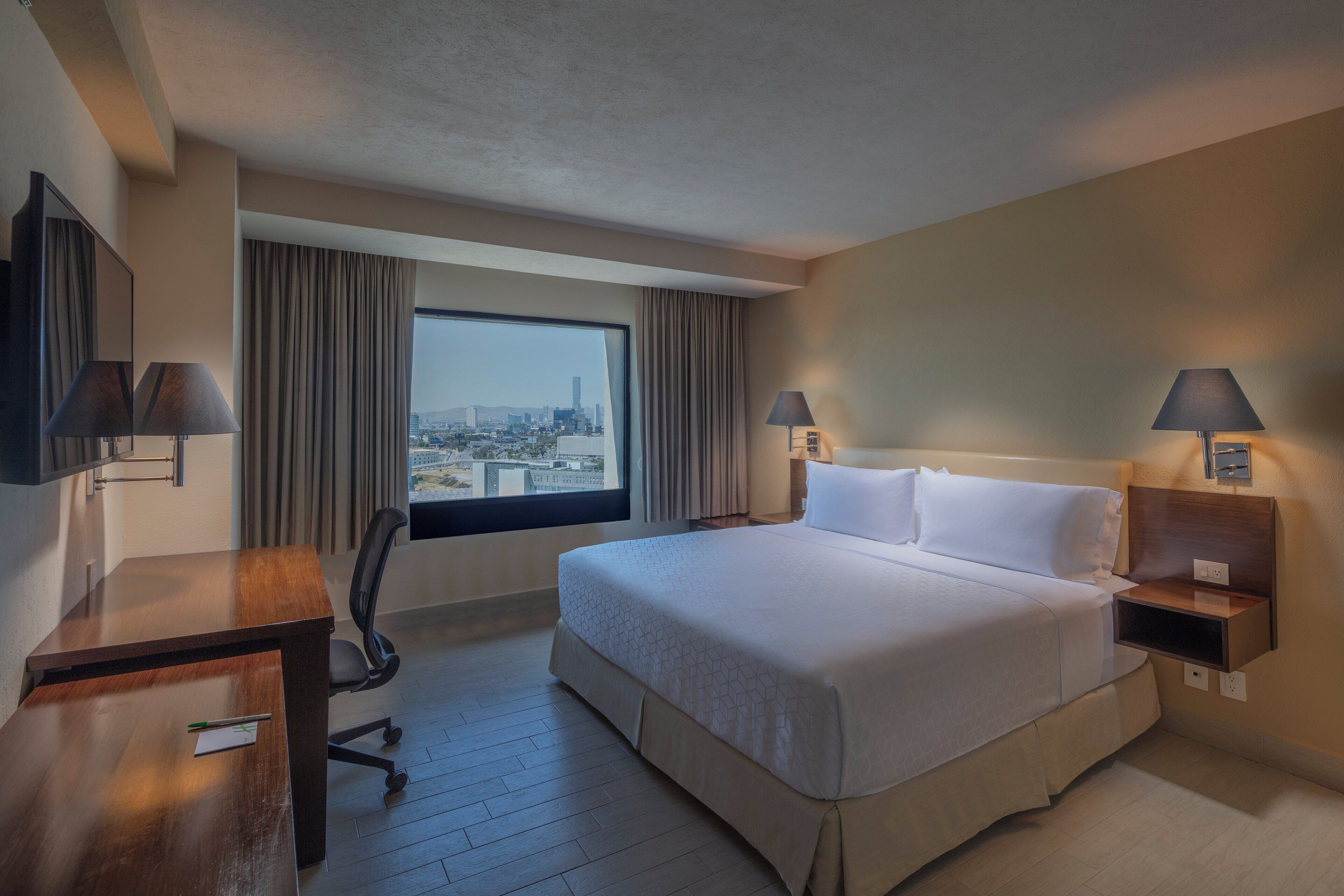 Habitación de hotel con una cama grande, un escritorio, dos lámparas, una silla y vistas a un paisaje urbano a través de un ventanal.