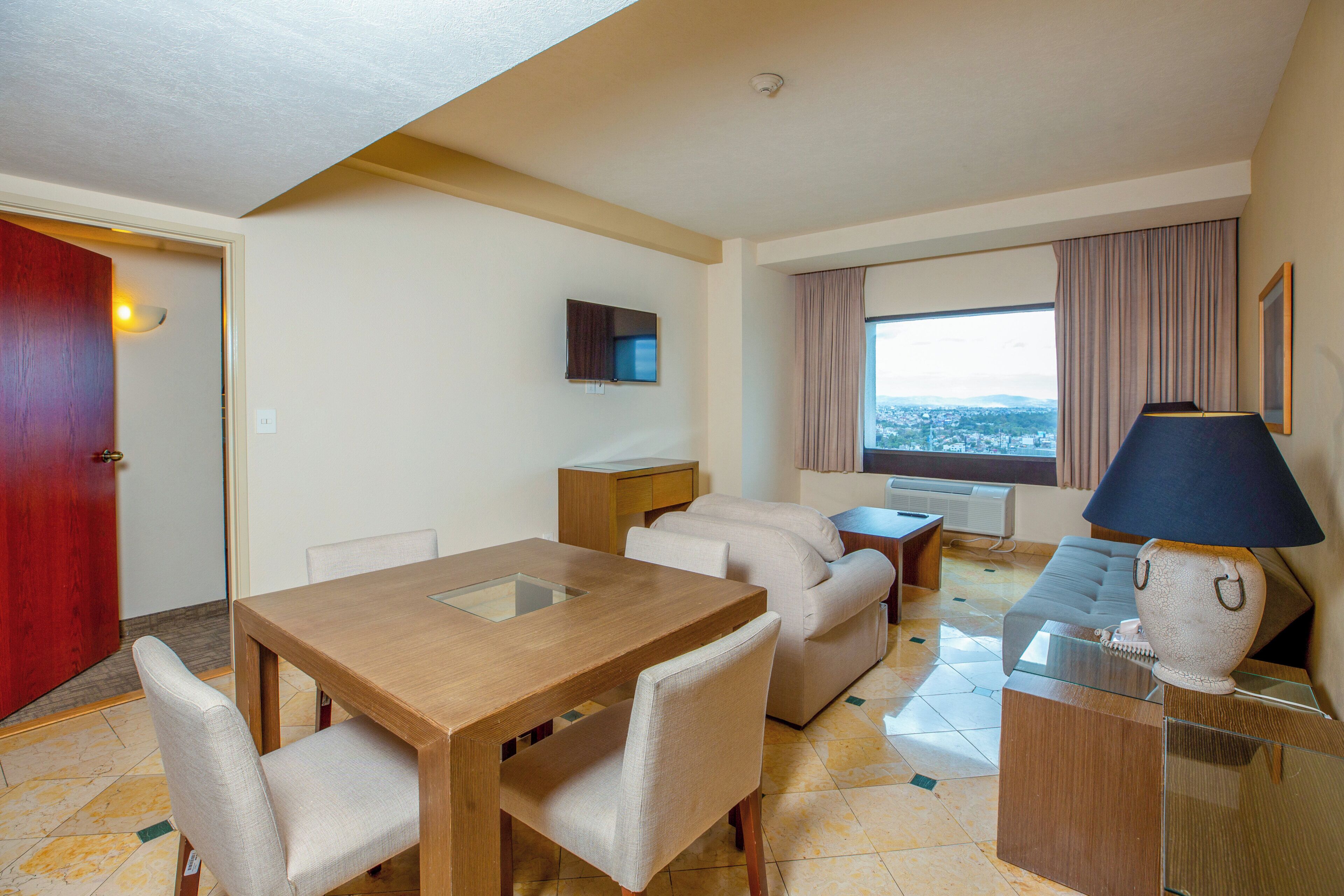 Habitación de hotel con mesa de comedor, sofá, televisor, lámpara y ventana con vistas.