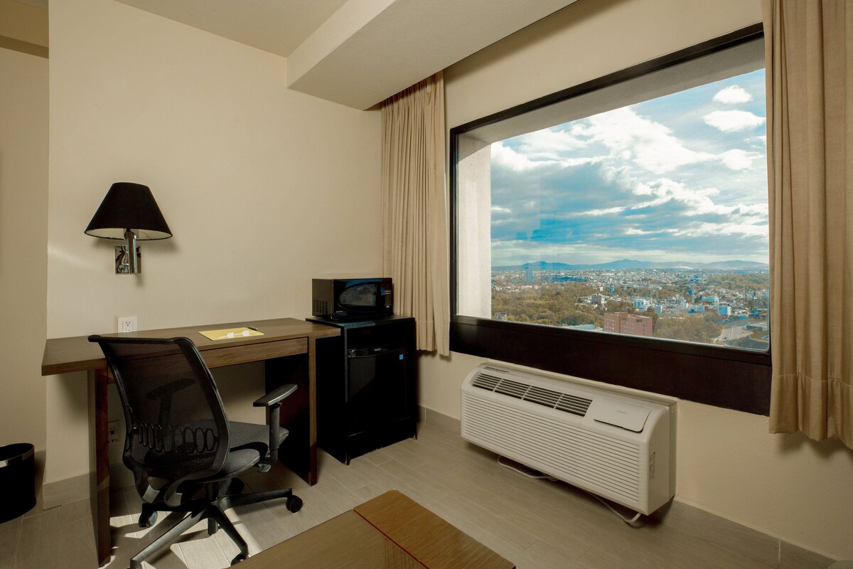 A hotel room with a desk, chair, TV, air conditioner, and a large window with a city view.