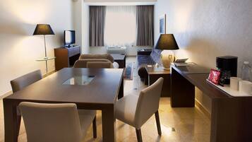 Une chambre d’hôtel équipée d’une table à manger, de chaises, d’un téléviseur, d’une lampe, d’une machine à café et d’une fenêtre avec des rideaux.