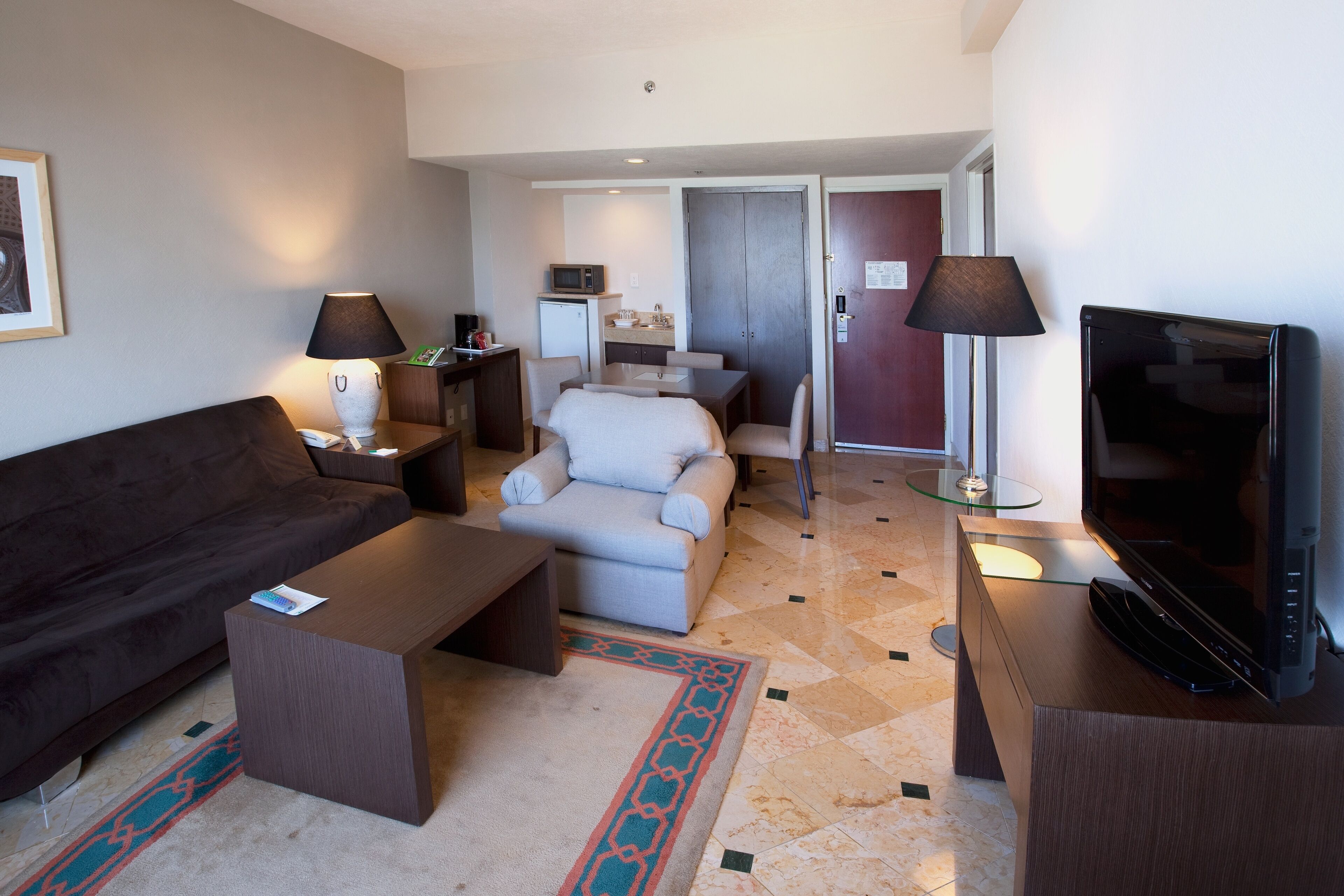 Una habitación de hotel moderna con un sofá de color oscuro, una pequeña mesa de centro, una televisión y un área de comedor con una mesa y sillas.