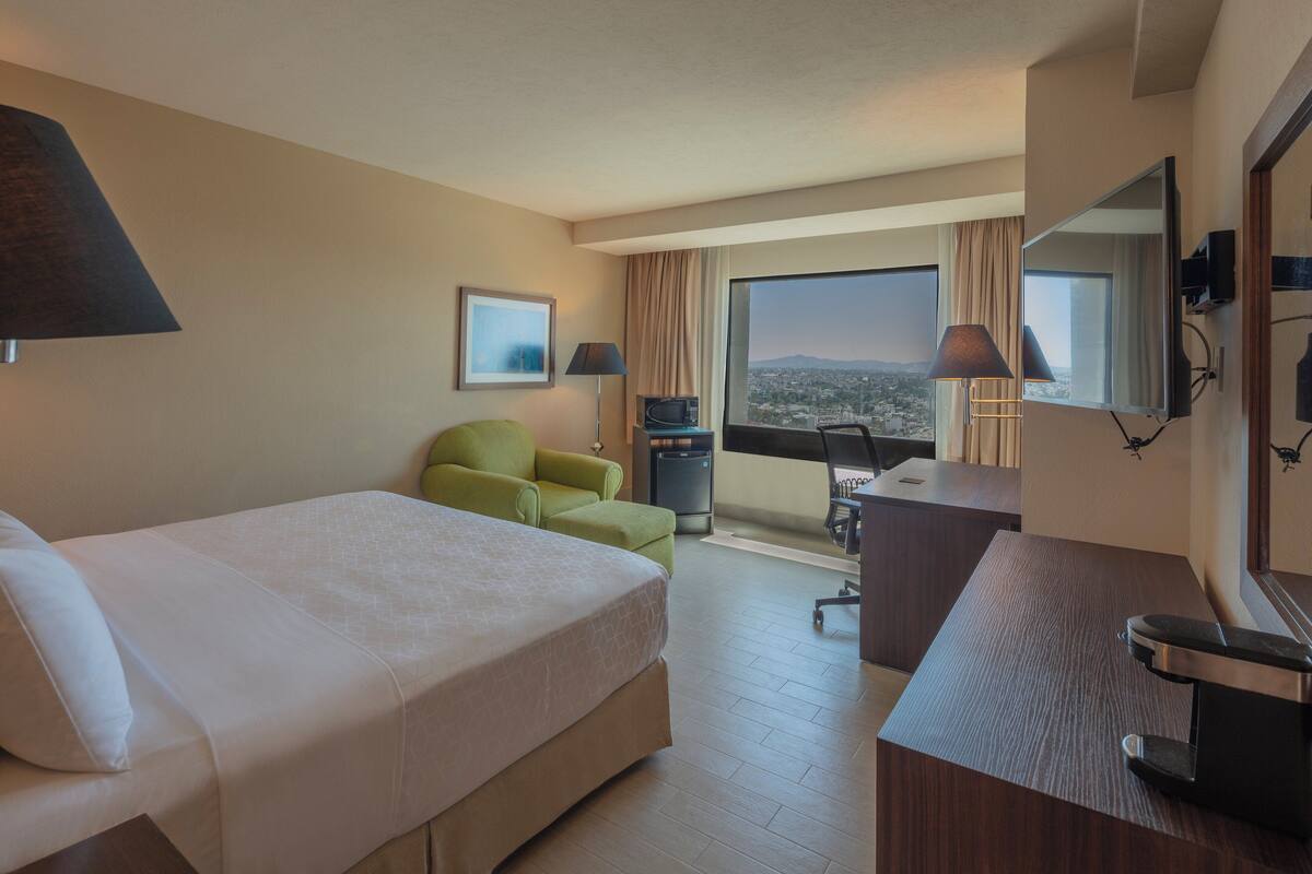 A hotel room with a bed, a desk, a chair, a TV, and a window with a city view.