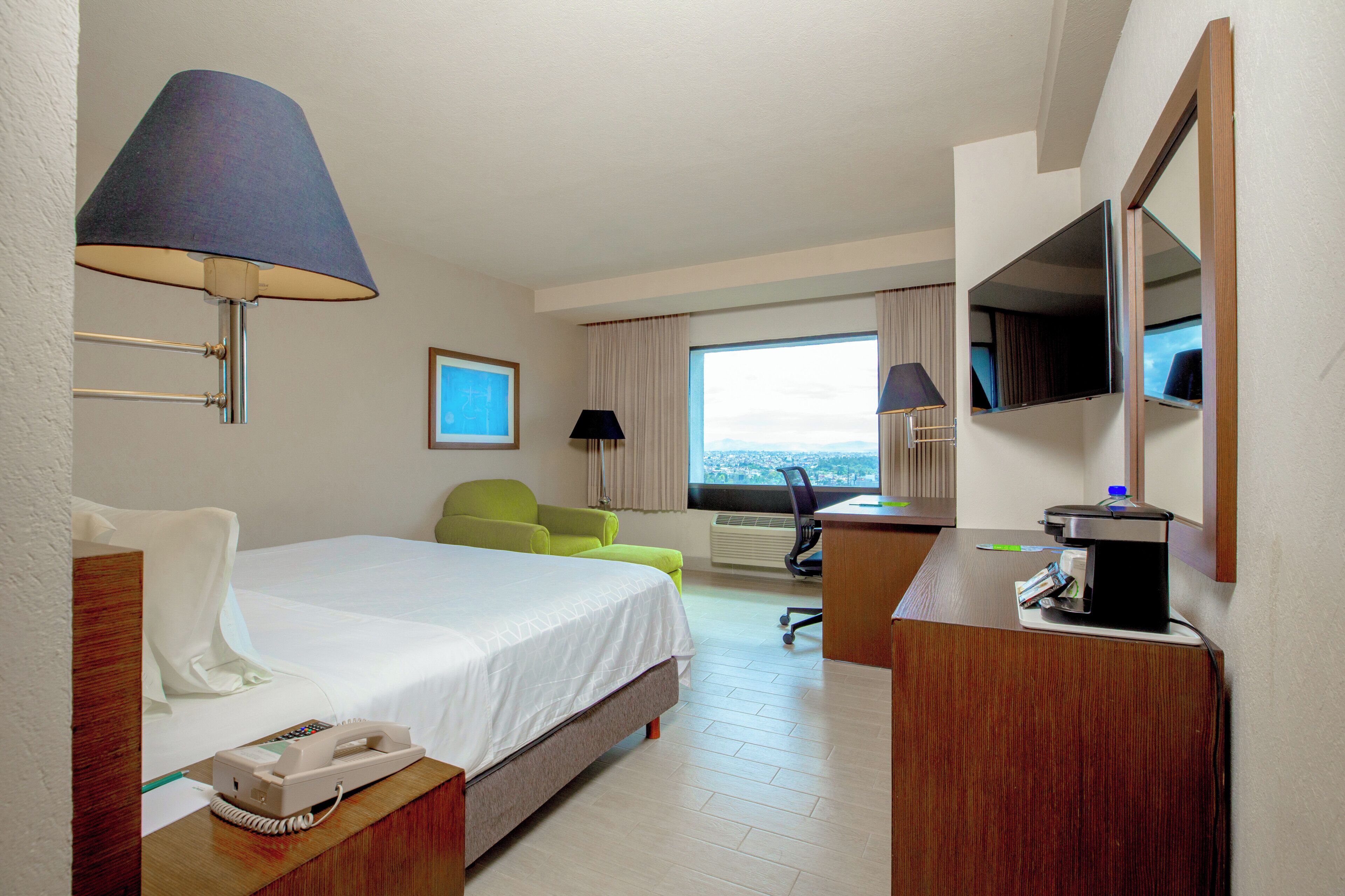Ein Hotelzimmer mit Bett, Schreibtisch, Stuhl, Lampe, Fernseher und einem Fenster mit Blick.