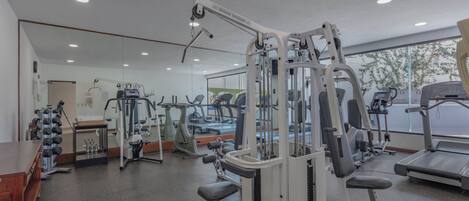 Un gimnasio con diversas máquinas de ejercicio, un espejo grande y una ventana que da al exterior.