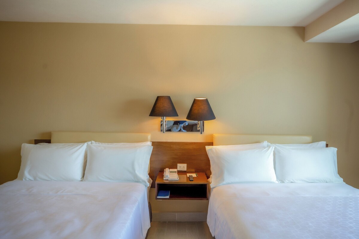 A hotel room with two beds, a nightstand with a lamp, a phone, and a remote control.