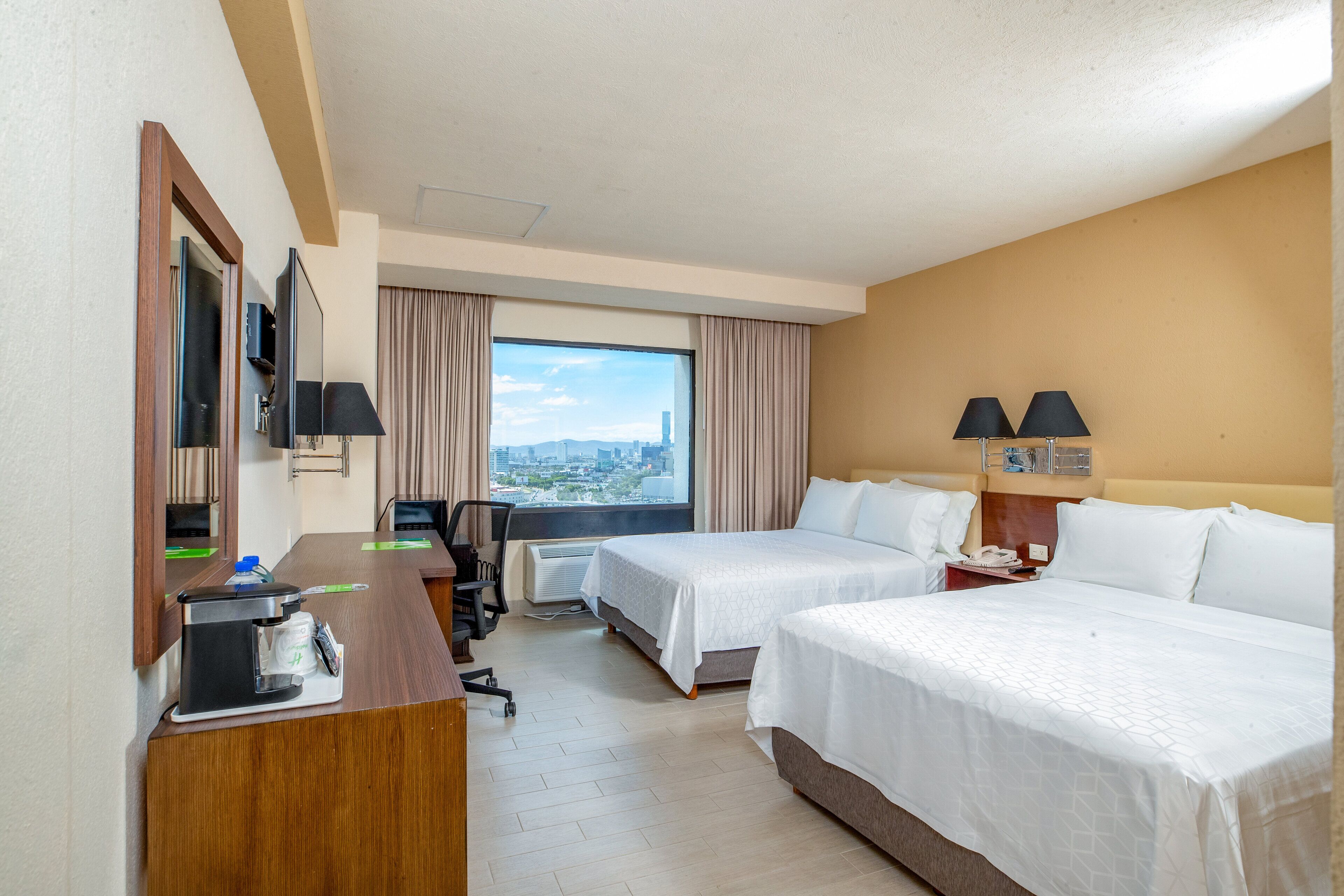 A hotel room with two beds, a desk with a computer, a chair, a coffee maker, and a view of the city.