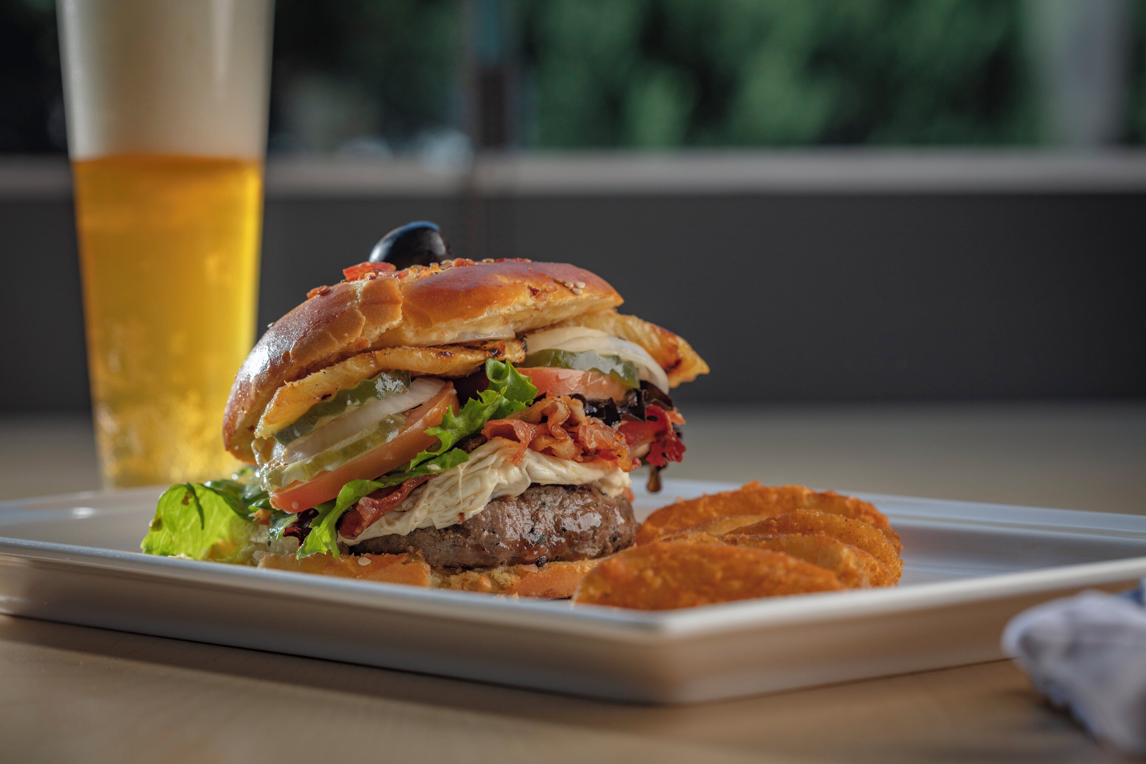 A burger with lettuce, tomato, and bacon on a white plate, accompanied by a glass of beer and a side of fries.
