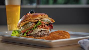 A burger with lettuce, tomato, and bacon on a white plate, accompanied by a glass of beer and a side of fries.
