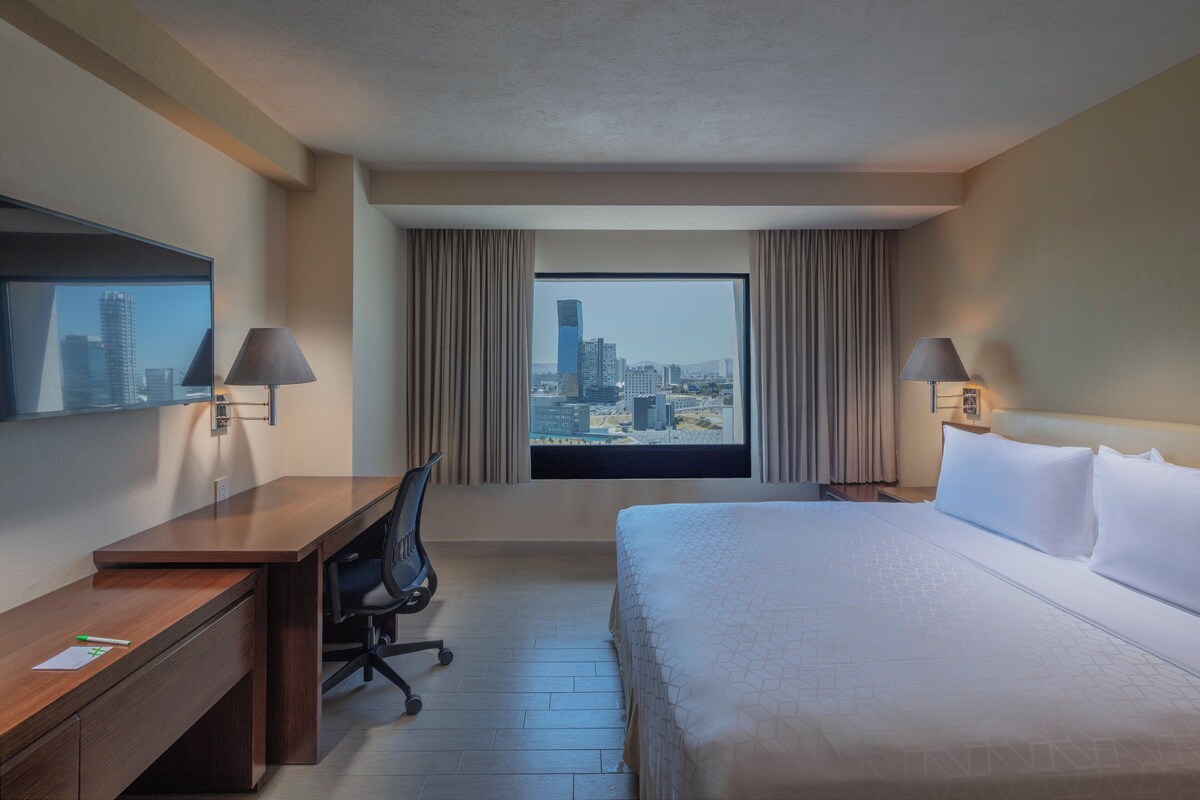 A hotel room with a large bed, a desk, a chair, and a view of a cityscape through a window.