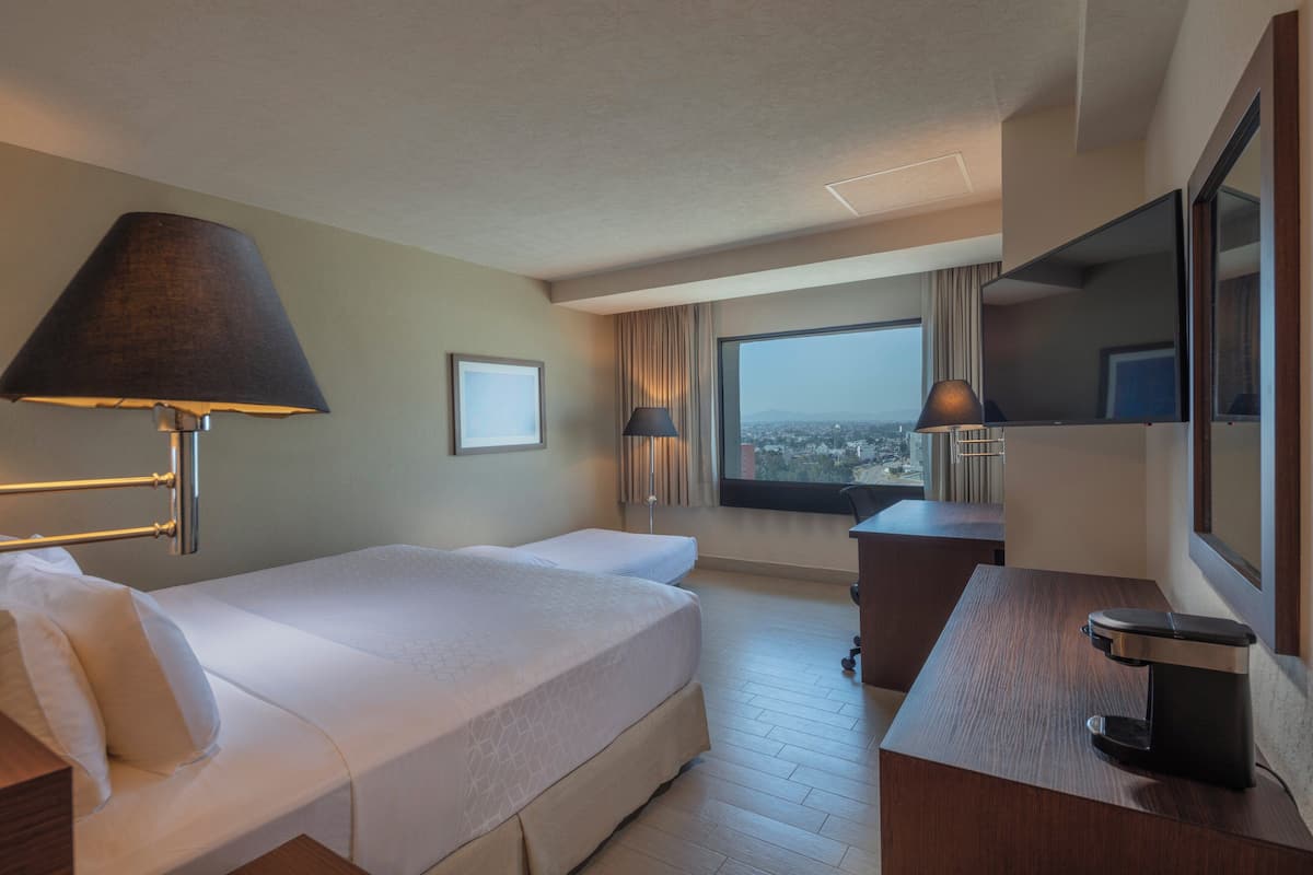 A hotel room with a bed, desk, and a large window offering a city view.