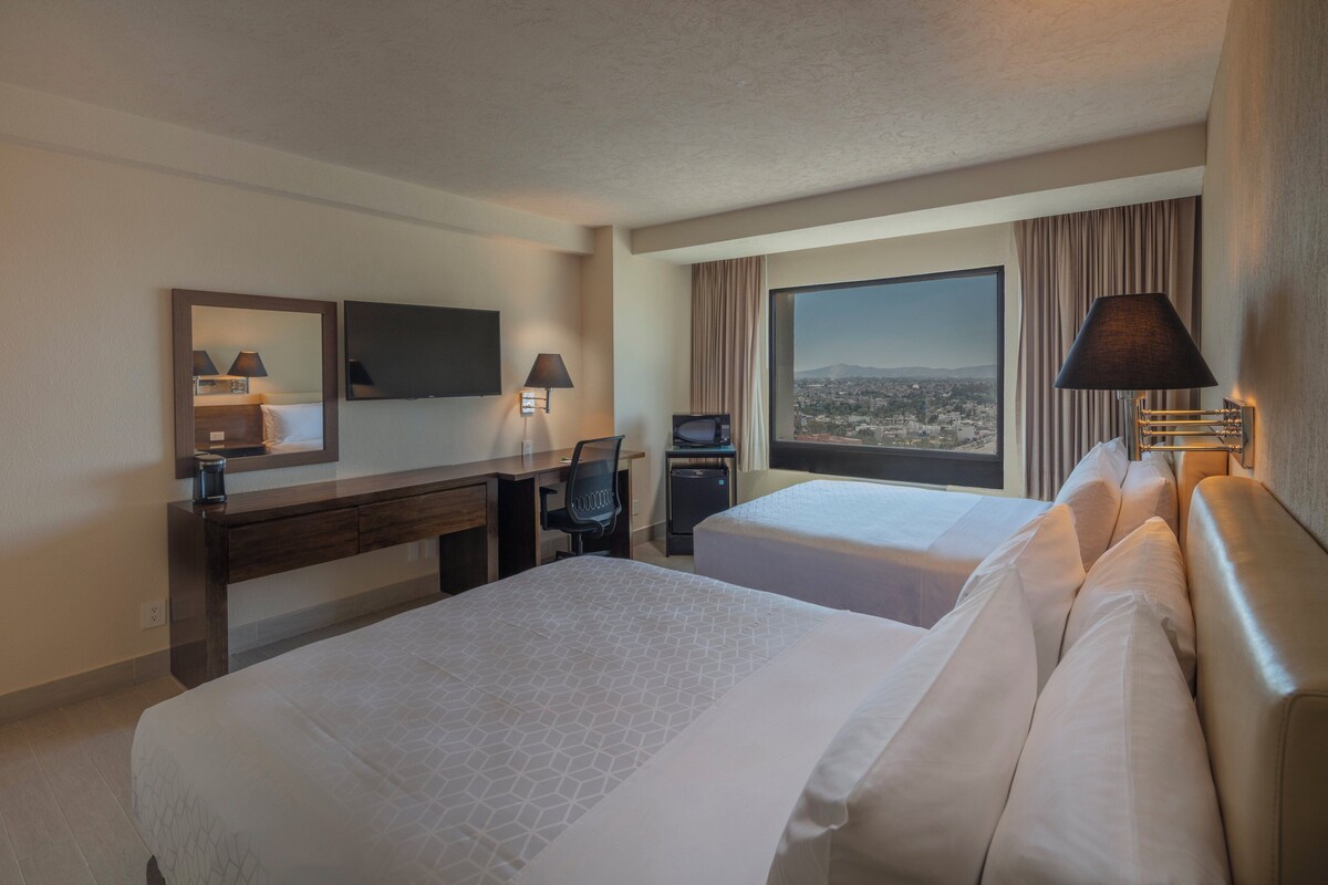 A hotel room with two beds, a desk, a chair, a TV, and a large window with a city view.