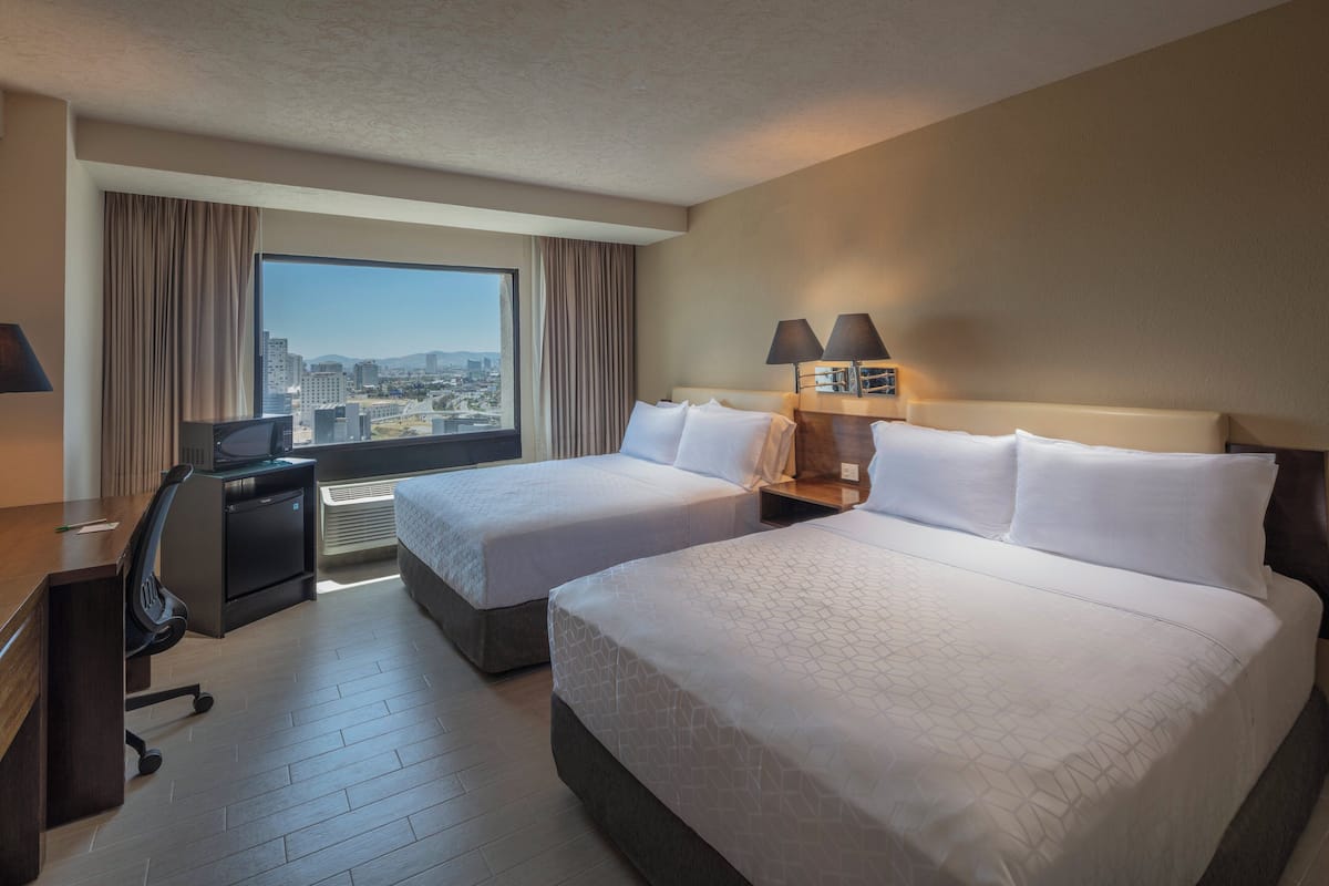 A hotel room with two beds, a desk, a chair, a TV, and a large window with a city view.