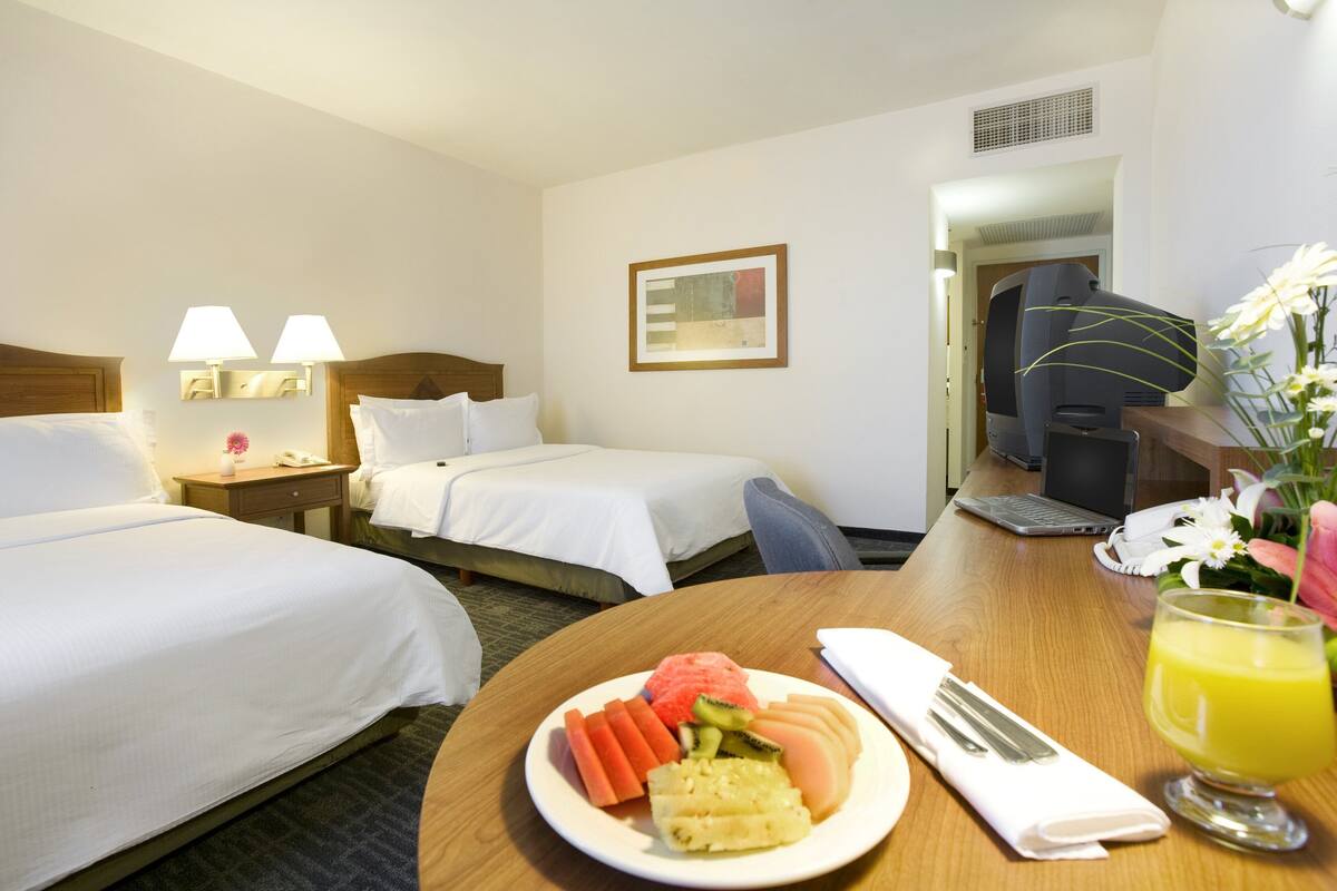 Une chambre d’hôtel avec deux lits, un bureau avec un ordinateur portable, une assiette de fruits et un verre de jus d’orange.