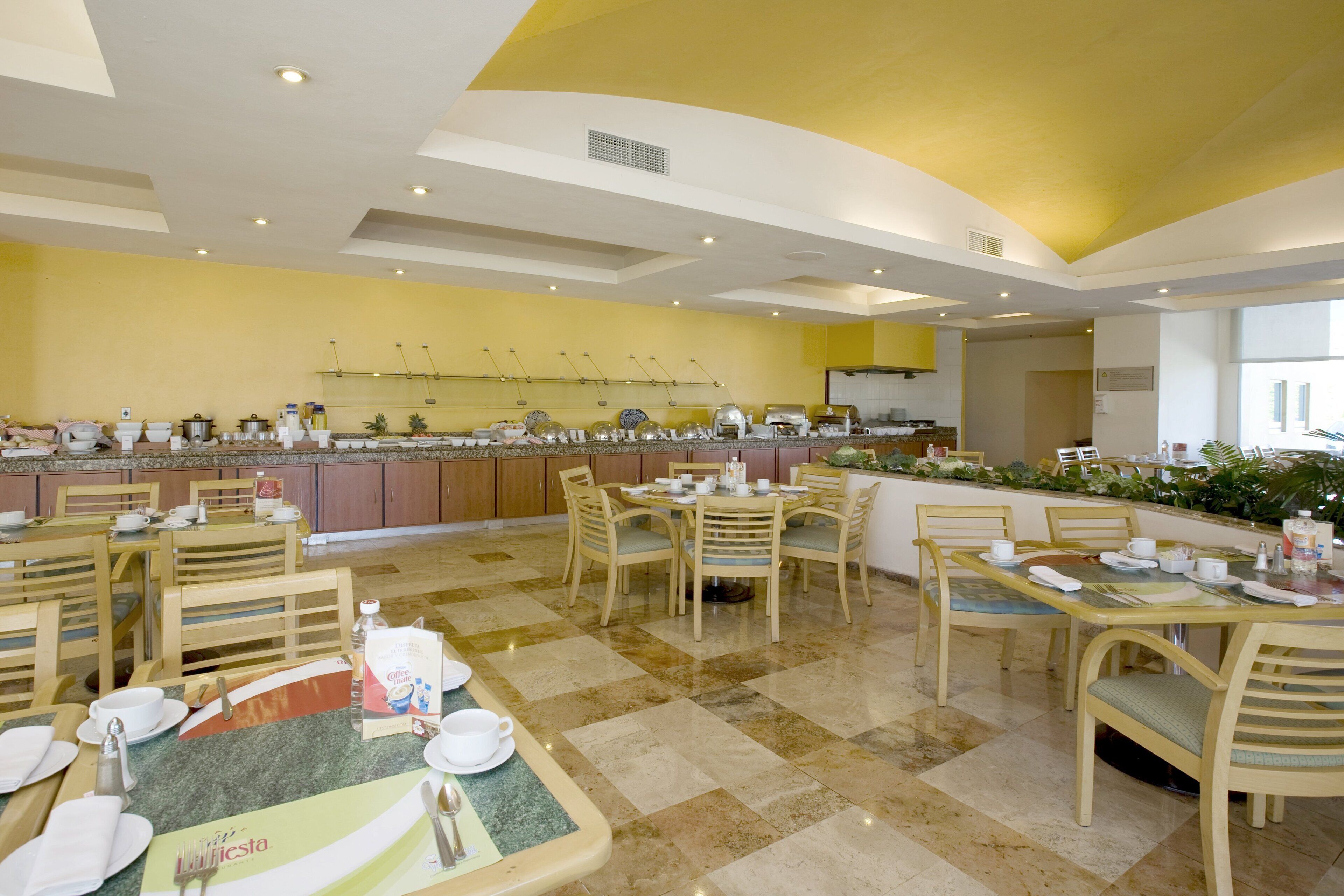 A spacious dining area with multiple tables and chairs, a buffet counter with various dishes, and a bright, well-lit ceiling.