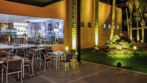 An outdoor dining area with tables and chairs, a covered seating area, and a well-lit garden with palm trees.