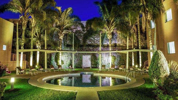 A pool area with lounge chairs and palm trees, surrounded by a building with a green wall and a staircase.