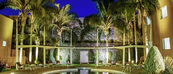 Zona de piscina con sillas de descanso y palmeras, rodeada por un edificio con pared verde y una escalera.