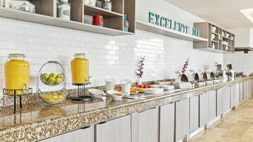 A kitchen with white tiled backsplash, a long counter with various food items, and a sign that reads "EXCELENTE DIA".